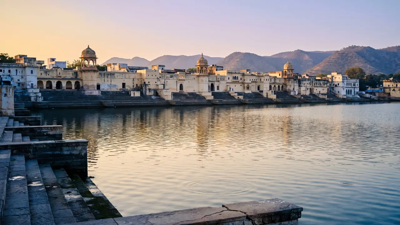 A long, low waterfront of pale plastered buildings and domed pavilions above lake steps at dawn, its reflection broken on rippled water, with layered hills behind.